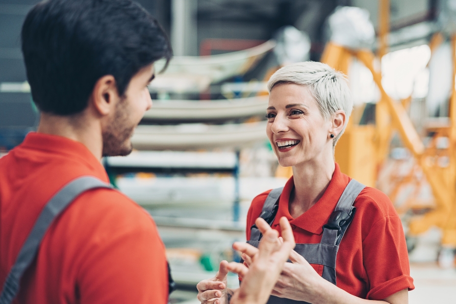 Man and woman in orange shirt and overalls conversing in manufacturing plant