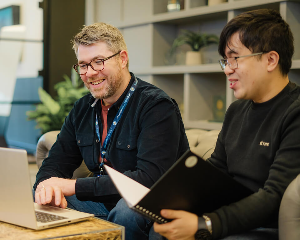 Two colleagues collaborate around a table with a laptop in a modern office setting.