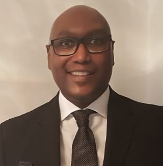 Man in black suit smiling in front of white wall.