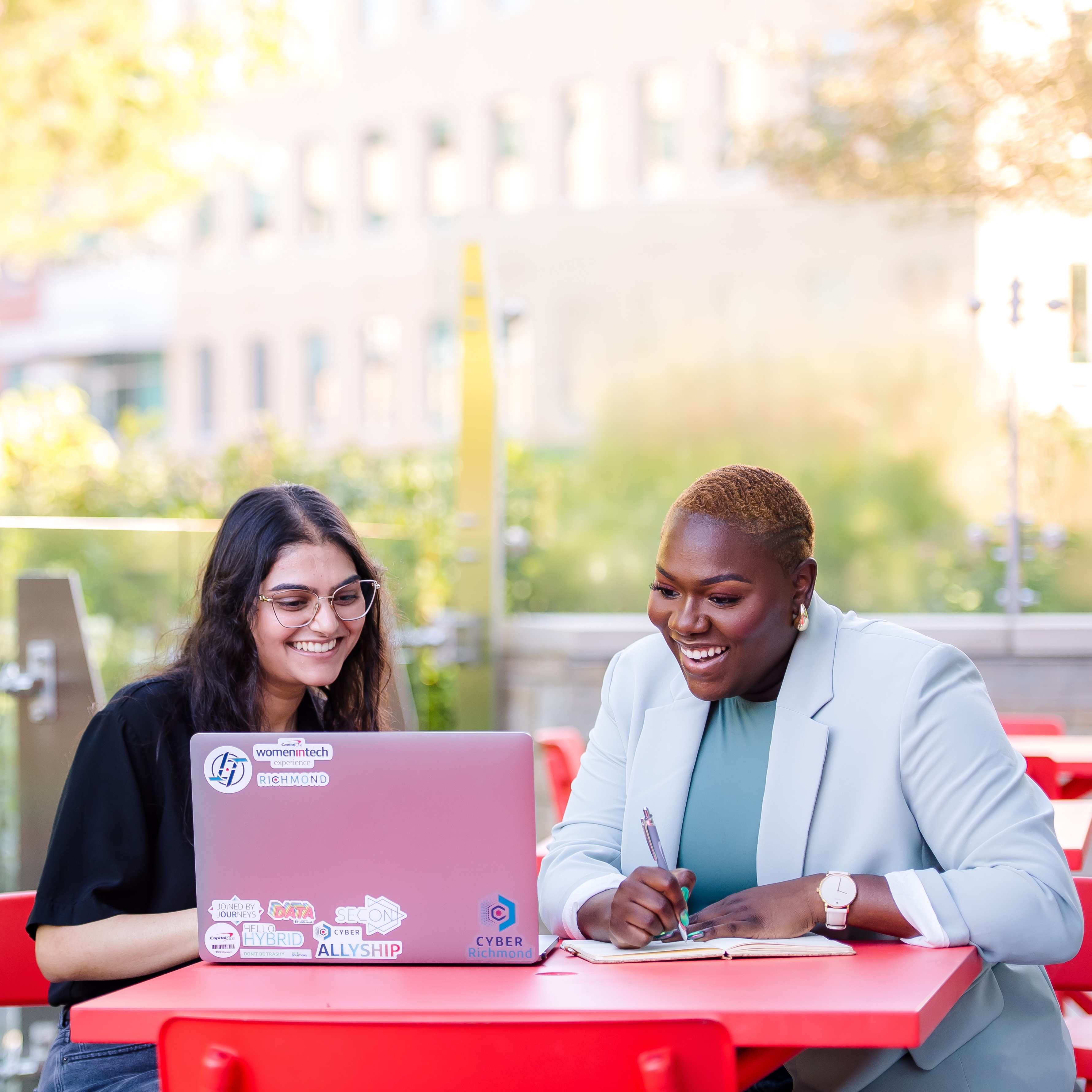 Two Capital One associates sit outside at a red table and look at a laptop together