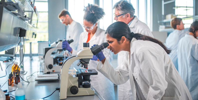 Researchers in lab coats working at microscopes in a laboratory with shelves of equipment in the background. Faces are anonymized.