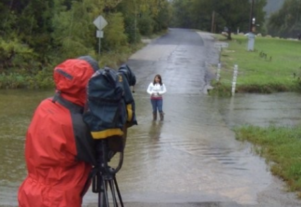 Camila stands in a road being recorded by a news camera.