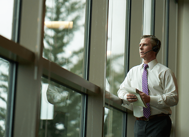 A man stands and looks out the window while talking on a telephone headset