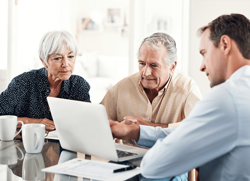 Senior man and woman look at a laptop, assisted by a younger man.