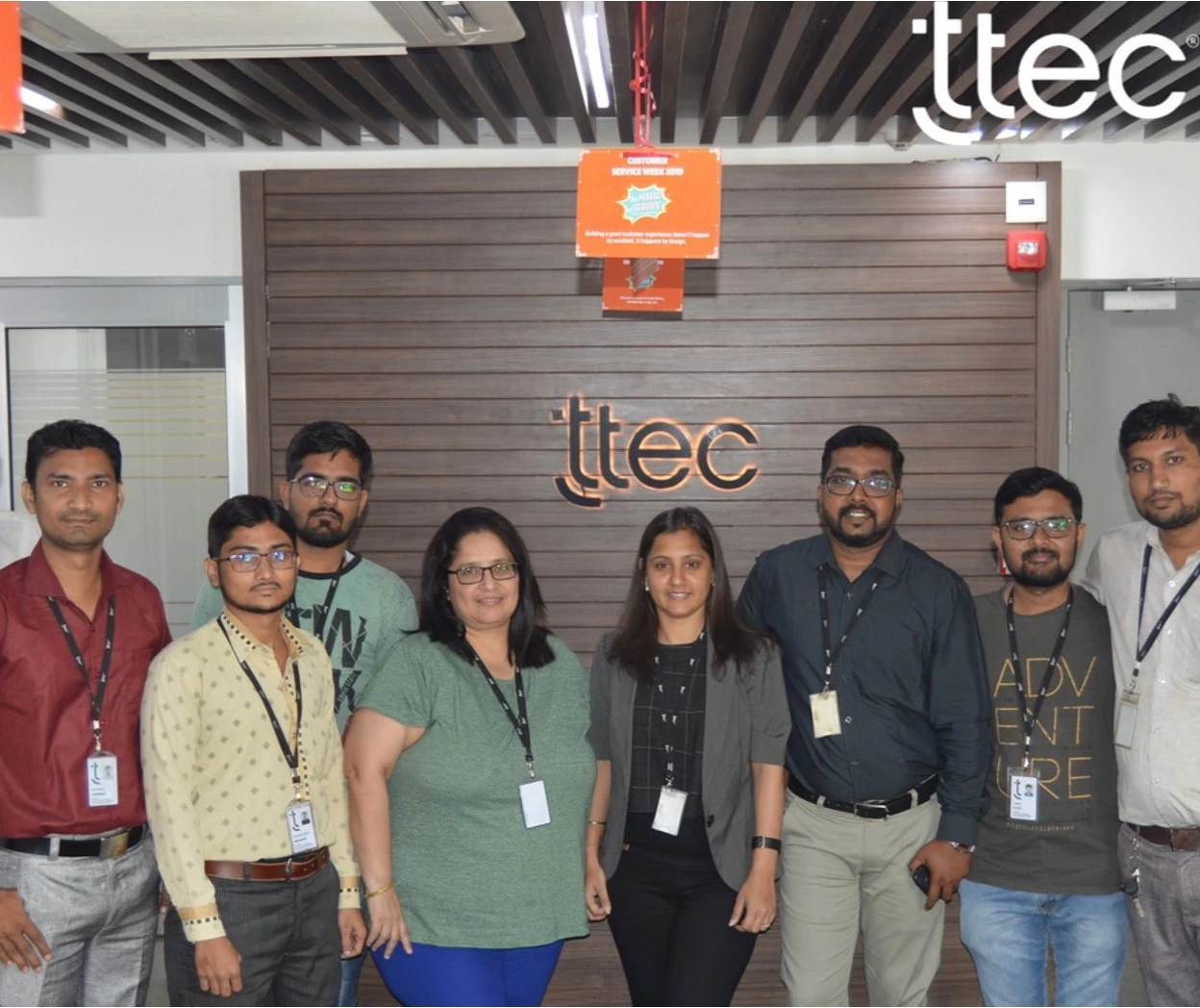A group of eight people, wearing office attire and ID badges, stand together in front of a ttec office sign on a wooden wall.