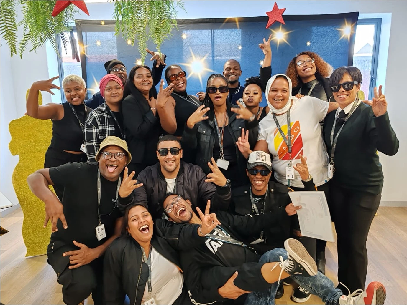 Group of coworkers posing joyfully indoors with peace signs and playful expressions, dressed in mostly black attire and sunglasses, under festive decorations with stars and greenery.