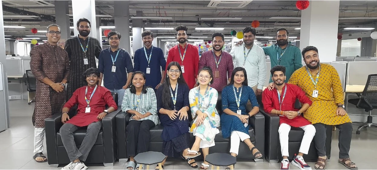 Office team posing together in traditional Indian attire, smiling in a modern open-plan workspace decorated with colorful hanging ornaments.