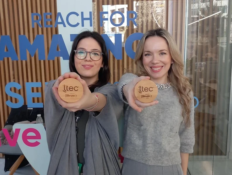 Two TTEC female employees holding up a company swag and smiling at the camera.