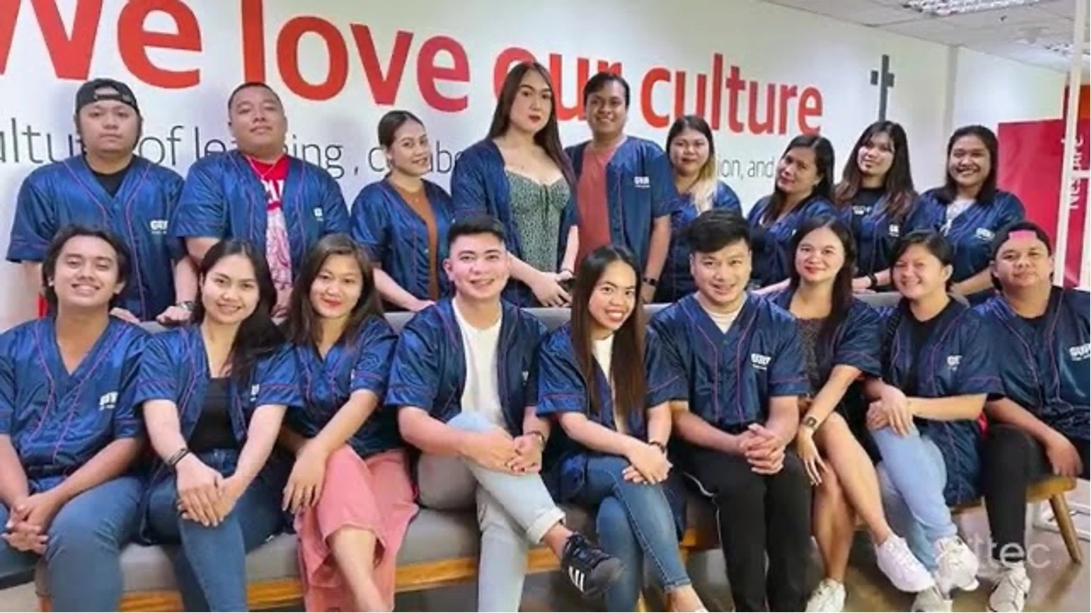 Group of employees wearing matching blue uniforms posing together in front of a wall with the text 'We love our culture'