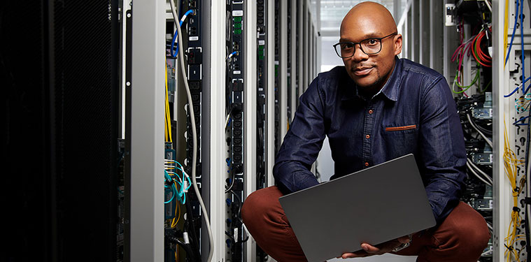 black man wearing glasses and collared shirt, squatting on the floor of a large server room while holding a laptop
