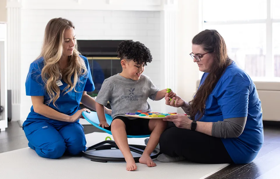 Two COFK nurses sit on the floor with a child seated on a cushion.