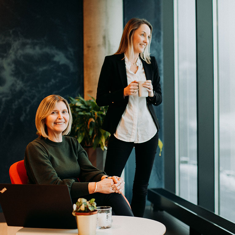 Two women drinking coffee next to an office window
