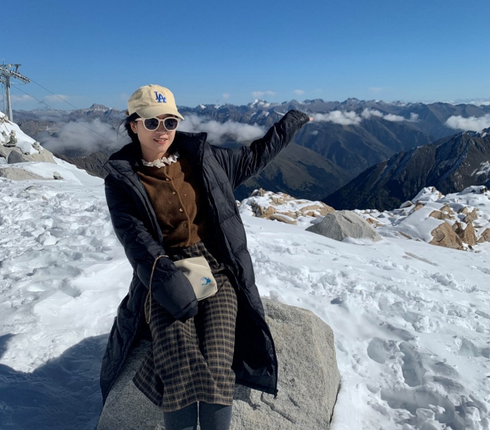 在一座覆盖着白雪的山上,一位女士戴着一顶鸭舌帽和墨镜,坐在一块大石头上