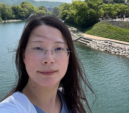 A woman with black long hair is standing in front of a river.