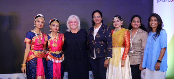 Group of seven women at an event, including two in traditional Indian dance costumes, standing together in front of a presentation backdrop