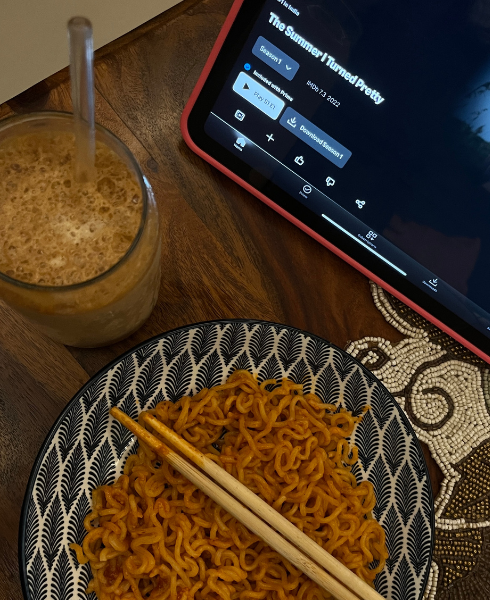 Bowl of noodles with chopsticks on a patterned plate, alongside a glass of coffee and a tablet displaying a media player on a wooden table.