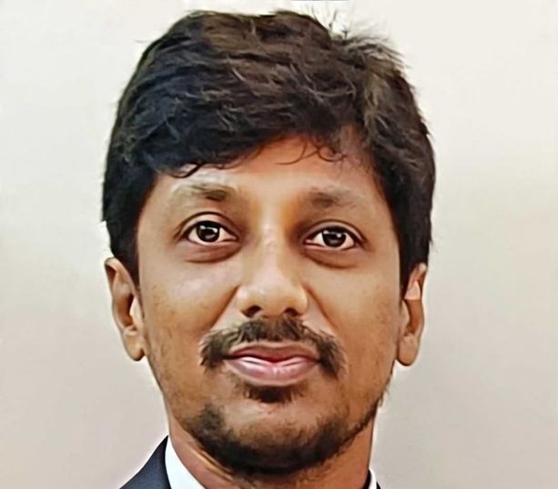 Ashok, is an Indian male, with dark hair with a side parting, dark eyes, and a neatly trimmed beard and moustache. He wears a dark suit and white shirt.
