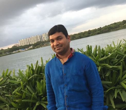A Man in blue shirt standing by a lake with green plants in foreground and city buildings in background under cloudy sky.