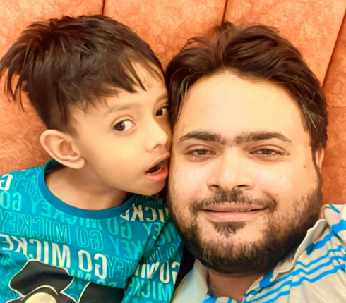 Shadaab and his son, both in blue and yellow tops. Shadaab has dark hair and a beard and is smiling to the camera.
