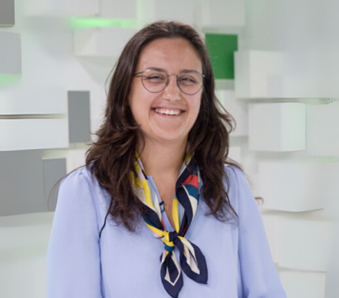 Simona, wearing a blue collared shirt and a multi-colored scarf, with brown long hair and glasses