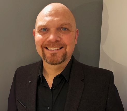 Tom, and English man, with a bald head and ginger beard, smiling warmly at the camera. He is wearing a black suit jacket and black button-up shirt against a gray background.