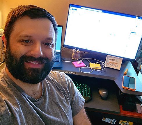 Reyad, a male Greek Lebanese American, is taking a selfie in his home office, smiling at the camera. He is at a desk with one computer monitor and a laptop. Workspace includes keyboard, mouse, and decorative items. Warm lighting creates a cozy atmosphere.