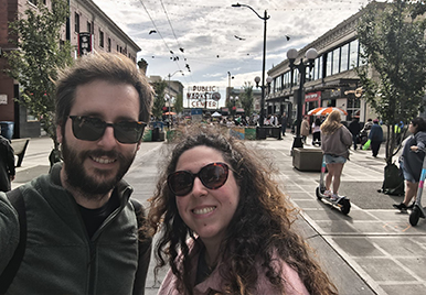 Andrea and her partner on an urban shopping street with pedestrians and storefronts under a cloudy sky.