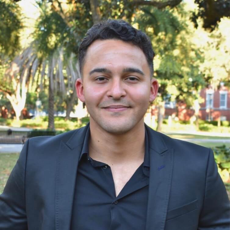 Diego, a male Hispanic Venezuelan, is standing in a park, he has dark short hair and wears a black suit with a black shirt.