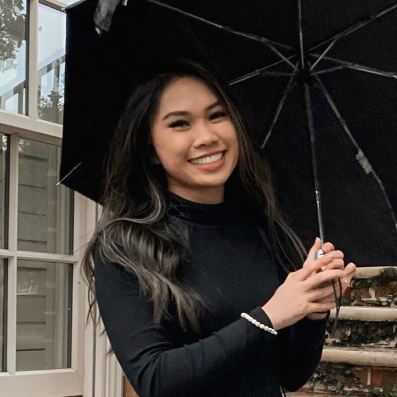APEX CRA participant Mimi Nguyen is holding an umbrella with both hands and smiling at the camera. She has dark long straight hair and wears a black pullover,