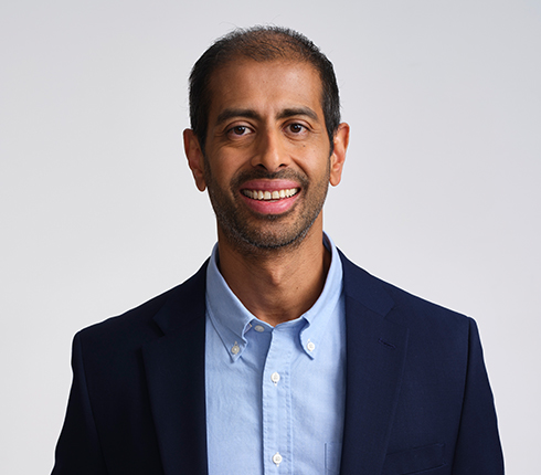 Chirag, an Indian man, smiling at the camera. He has short dark hair and is wearing a navy blue blazer and light blue button-down shirt against. The background is light gray.