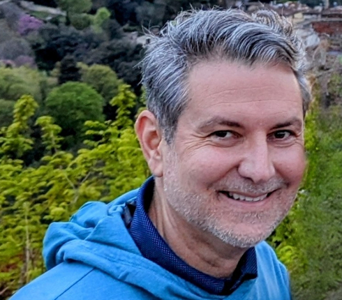 Chris, white male, with silver grey hair, some stubble, smiling towards the camera, wearing a blue hooded jacket