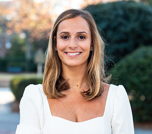 Lillie, a white American, with shoulder-length brown hair and she is wearing a white outfit. It's a portrait picture of her smiling at the camera, against a blurred background of green shrubs and autumn foliage.