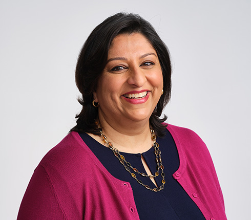 Professional headshot of a Hina, a female American, she is smiling with a warm, friendly expression. She has dark hair, wears a vibrant pink cardigan over a navy top and gold jewelry.