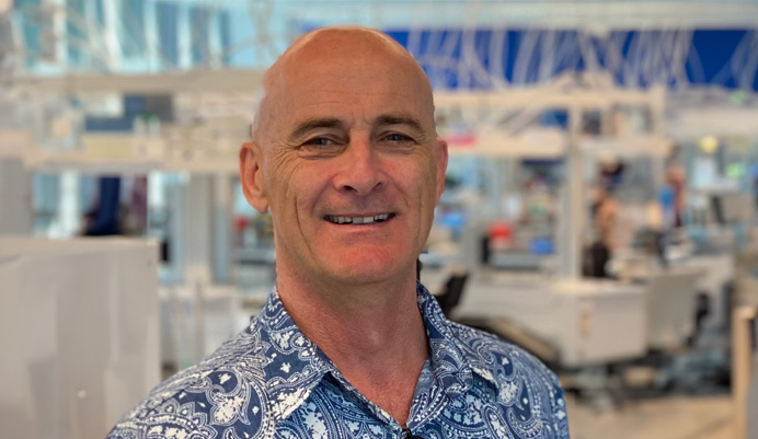 Lachy, a white man in blue patterned shirt stands in office smiling. He appears friendly and professional in a casual business environment.
