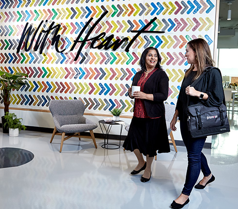 Colorful chevron wall art with "With Heart" text. Two women walking and talking in modern office space. Gray chair, small table, and plants visible. Bright, open interior with polished floor.