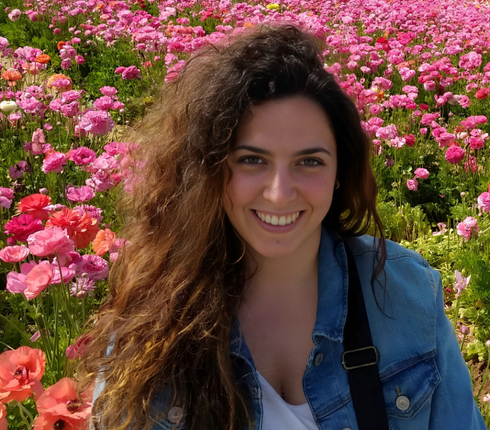 Andrea, a Spanish woman, with long curly hair smiles brightly. She is wearing a denim jacket. She's surrounded by a vibrant field of pink and red flowers in full bloom, creating a colorful, cheerful backdrop.