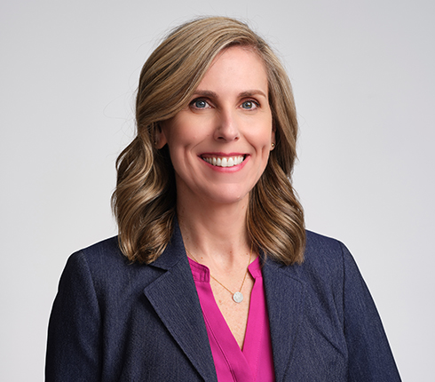 Stacy Hurt, a white female American who has shoulder-length wavy hair. She is wearing a navy blazer over a bright pink blouse, smiling at the camera, against a light gray background.