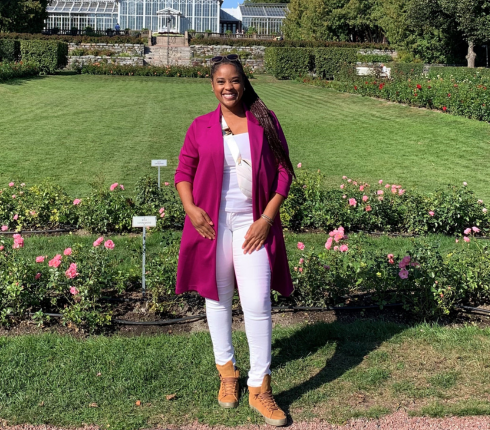 Yoela is standing in a garden setting, with flowers and grass around her. she has her hair tied back, and is wearing a pink jacket and a light coloured top and light trousers.