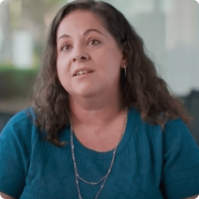 Screenshot of one of the employees speaking in the video. Cristina is a Senior Manager in Clinical Operations, and she is a white Italian American, she has curly long hair and wears a blue shirt.