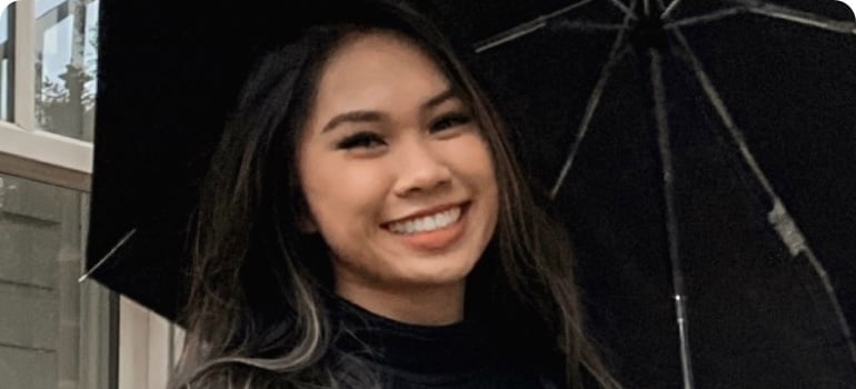 Mimi, a female Vietnamese American, is holding an umbrella with both hands and smiling at the camera. She has dark long straight hair and wears a black pullover,