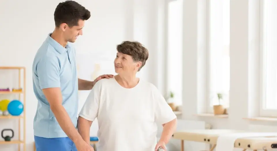 young male physical therapist assisting an older female patient in a bright, sunlit hospital room