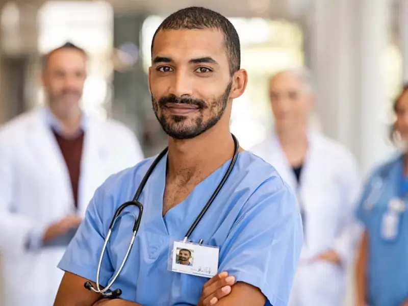 Man in blue scrubs stands in hallway with other medical professionals in the background