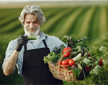 An older man with gray hair and beard, holding fresh produce in a field, smiling.