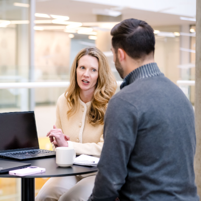A male and female colleague seated at a small table in front of a computer having a conversation.