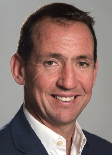 A professional headshot of a man, Tadhg O'Malley, smiling in a colored shirt and suit jacket