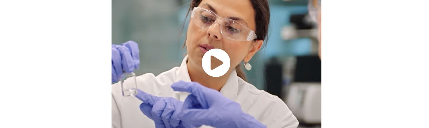 Woman in lab coat and glasses is wearing blue rubber gloves and looking closely at a test tube. Over the image is a white "play" button, indicating this is a video.