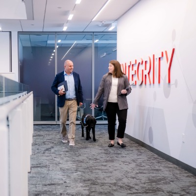 man and woman walking down an office hallway with a black service dog between then. Integrity is emblazoned on the wall.