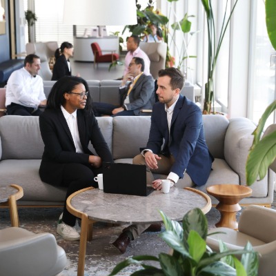Man and woman seated in a lobby talking with one other while looking to a computer that is placed between them