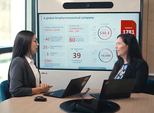 Watch video: 2 female coworkers conversing at a conference table in front of a smart board