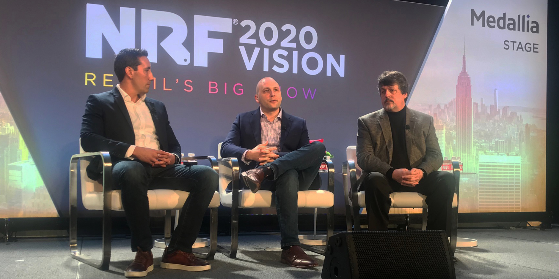 "Three men sit in chairs onstage in front of a big screen showing the NRF Big Show logo"